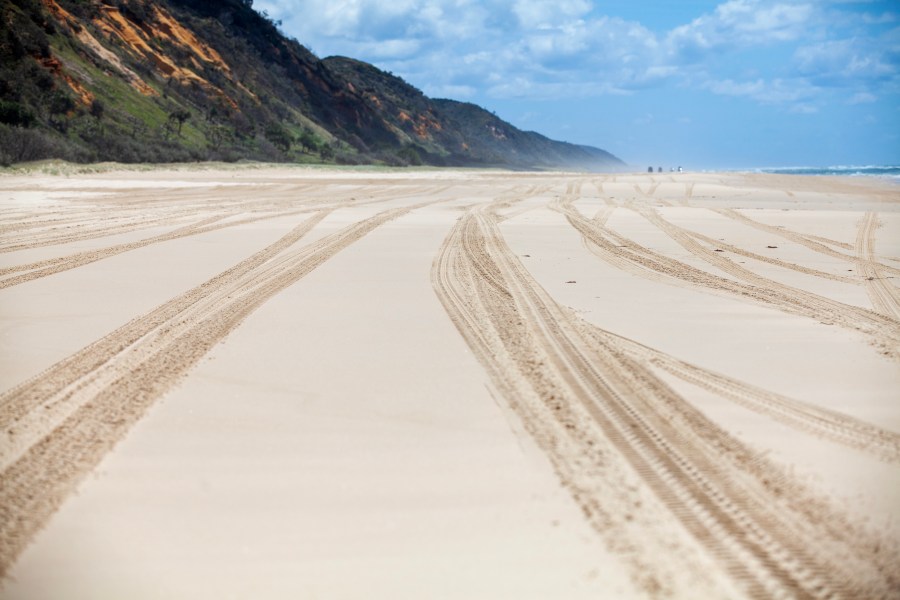 K'gari (formerly Fraser Island) driving along the beach in the main island highway. Cars in background. Wheel marks in sand. Cliffs on one side and glimpse of ocean across the wide beach on the right side.